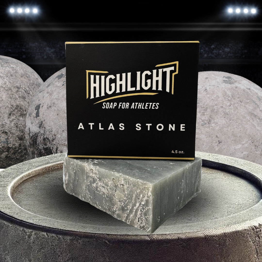 A black box of Highlight Soap for Athletes, labeled 'Atlas Stone,' is placed on a rugged, stone-like surface. In the background, large gray stones resembling atlas stones used in strength competitions are illuminated by stadium lights.