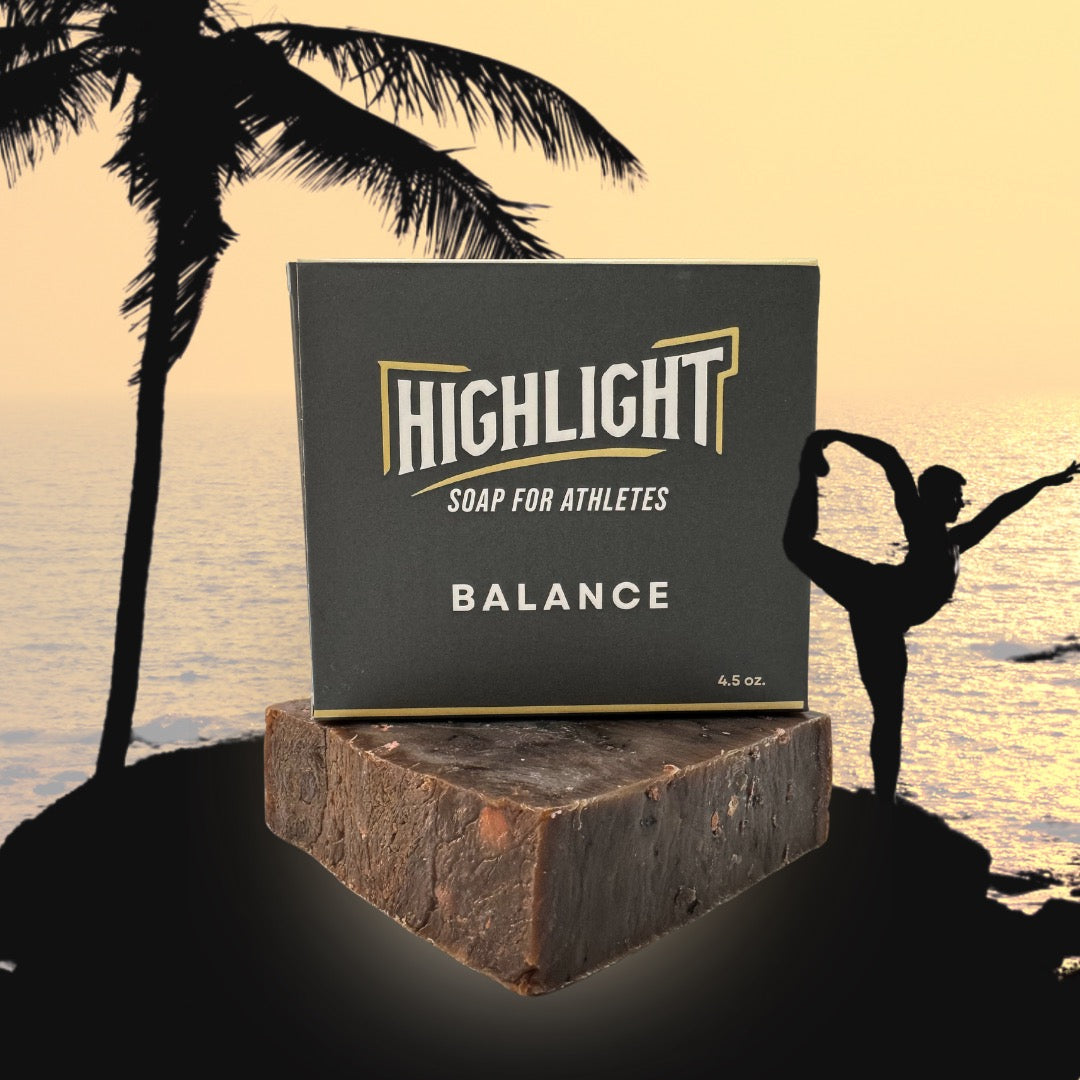 A grey box of Highlight Soap for Athletes, labeled 'Balance,' is set on top of a rustic soap bar. The background features a serene sunset over the ocean, with a silhouetted palm tree and a person gracefully balancing in a yoga pose, conveying a sense of harmony and tranquility.