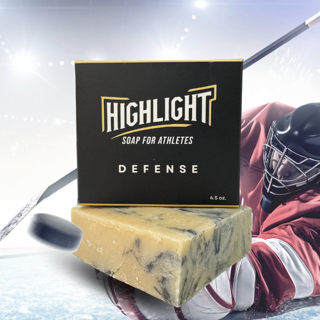 A black box of Highlight Soap for Athletes, labeled 'Defense,' rests on a marbled soap bar. The background shows a dynamic hockey scene, with a goalie in red gear making a save, a hockey stick, and a puck in motion, emphasizing the soap's strong and protective theme."