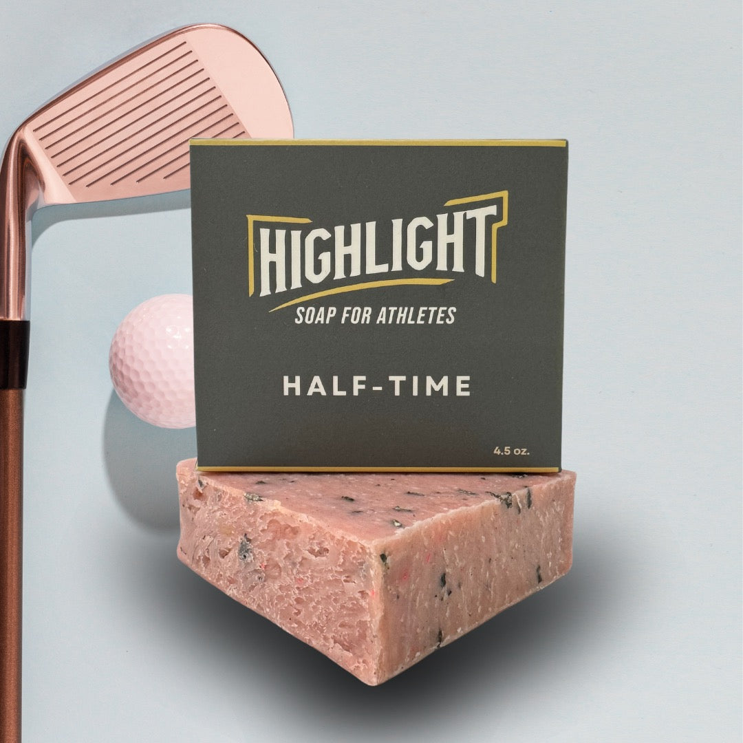 A grey box of Highlight Soap for Athletes, labeled 'Half-Time,' rests on a speckled, pink soap bar. In the background, a rose gold golf club and a white golf ball are positioned against a light blue backdrop, evoking a sporty and leisurely theme.