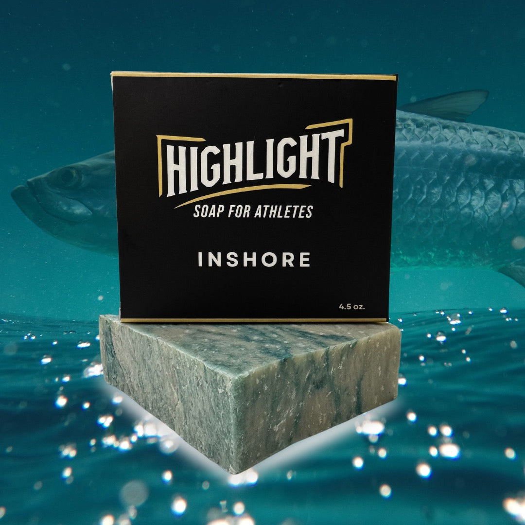 A black box of Highlight Soap for Athletes, labeled 'Inshore,' sits atop a marbled, greenish-blue soap bar. The background depicts an underwater scene with a large fish swimming by, enhancing the soap's coastal and aquatic theme.