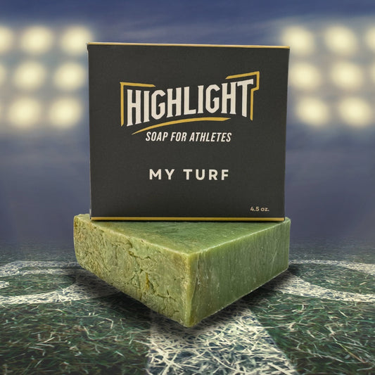 A black box of Highlight Soap for Athletes, labeled 'My Turf,' sits on a green soap bar with a natural, grassy texture. The background features a sports field under bright stadium lights, evoking an athletic and competitive atmosphere.